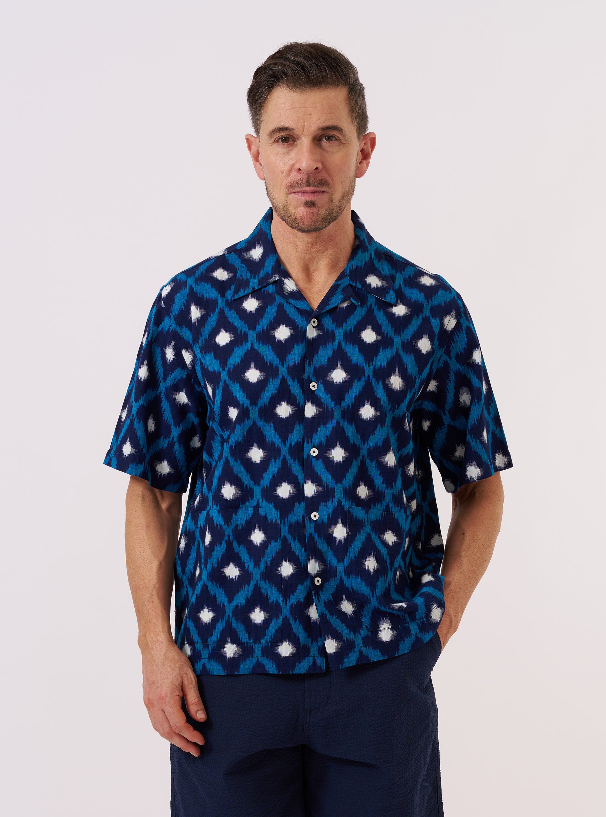 A model wears the Indigo Diamond Ikat Deck Shirt over a light-colored top, standing against a plain white background. The shirt features a distinctive three-color diamond ikat pattern in indigo, a relaxed wide fit, and a clean one-piece collar. Two large patch pockets are prominent on the lower front of the garment, which is shown with the top button undone and the short, loose sleeves visible.