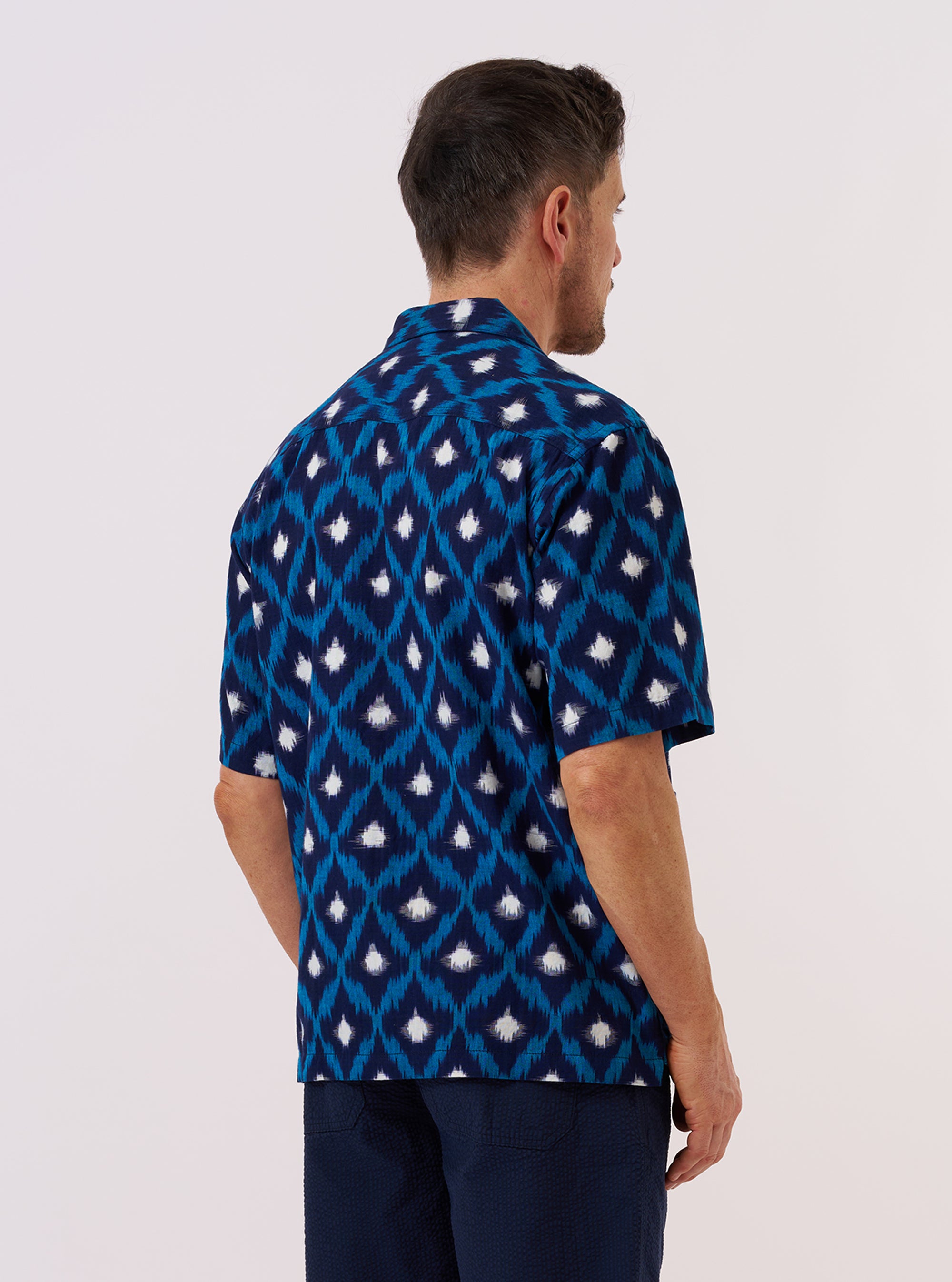 A model is wearing the Indigo Diamond Ikat Deck Shirt, shown from the back against a plain white background. The shirt features an all-over diamond ikat pattern in shades of indigo and white. The garment has a relaxed fit, a straight hem, and short sleeves, with a simple back yoke and a central vertical seam. No mannequin is present in the image.