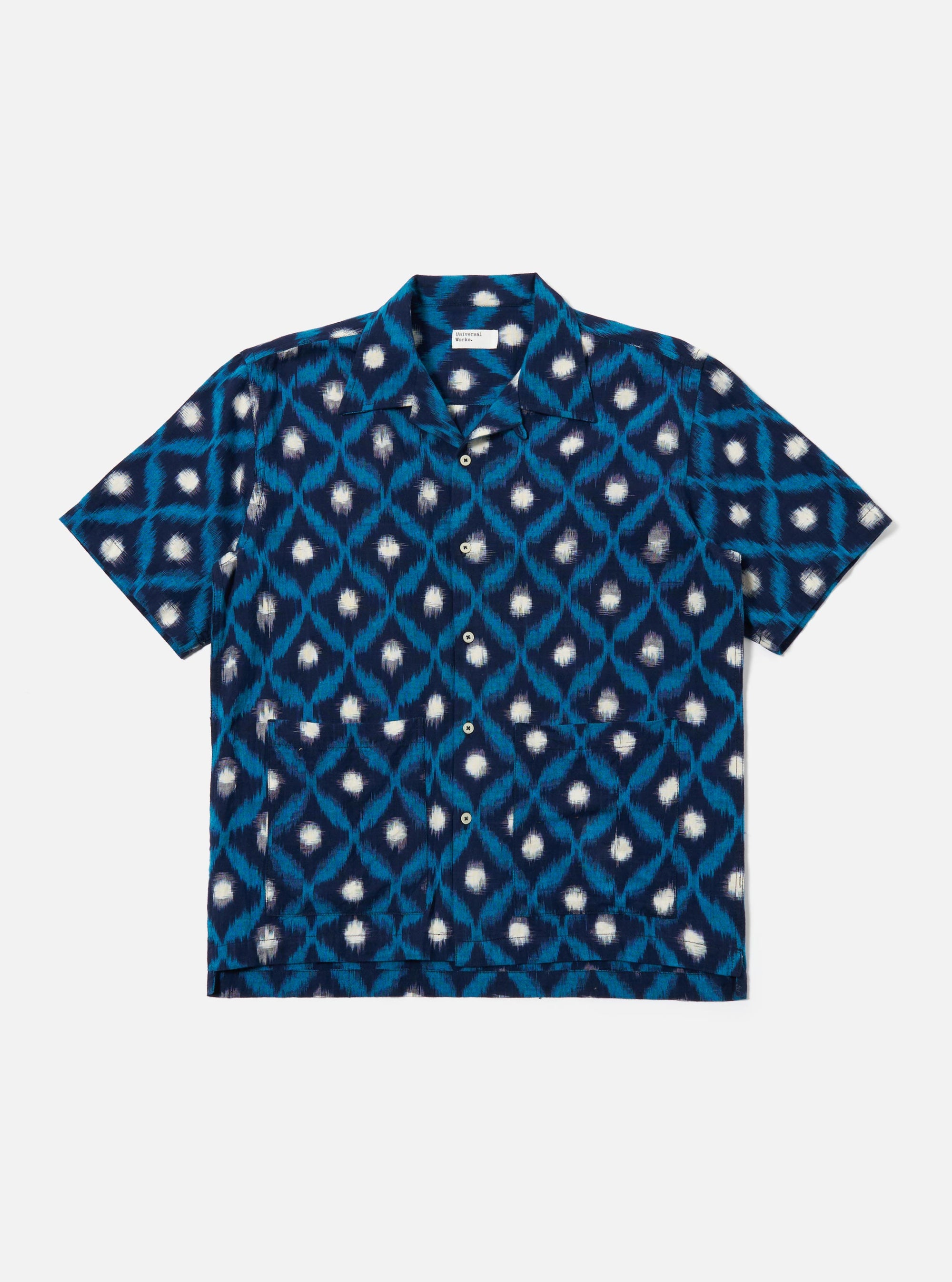 The Indigo Diamond Ikat Deck Shirt is presented as a flat lay against a white background. The shirt features a one-piece collar, short loose sleeves, and two large patch pockets at the lower front. The fabric is decorated with a three-color double ikat diamond pattern in indigo tones.