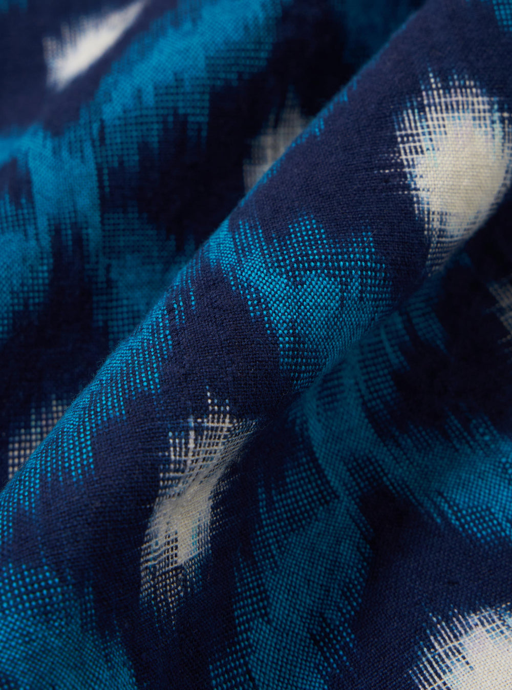 A close-up fabric swatch of the Indigo Diamond Ikat Deck Shirt is shown, highlighting the intricate diamond-shaped ikat pattern in shades of indigo and white. The textile features a visible woven texture characteristic of 100% cotton.