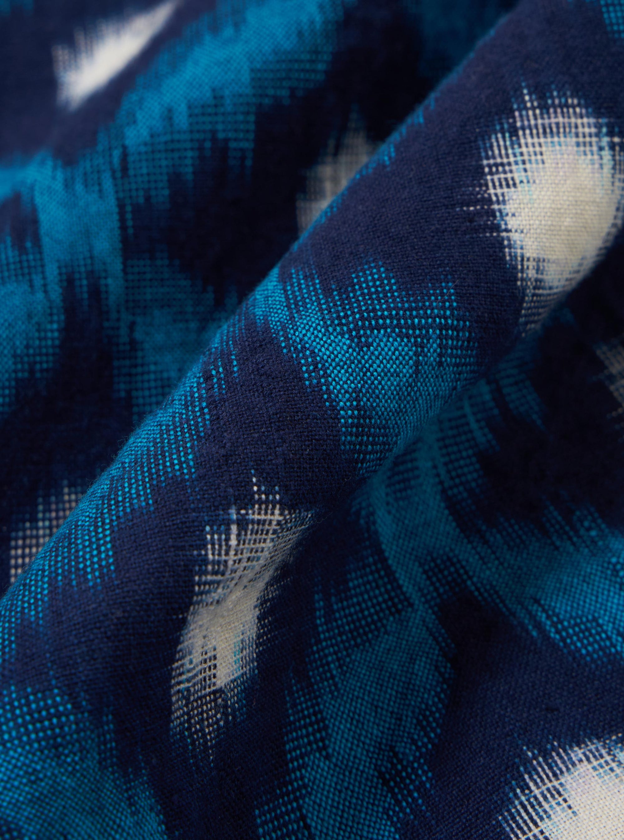 A close-up fabric swatch of the Indigo Diamond Ikat Deck Shirt is shown, highlighting the intricate diamond-shaped ikat pattern in shades of indigo and white. The textile features a visible woven texture characteristic of 100% cotton.