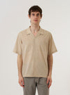 A model wears the Beige Fine Organic Cotton Sal Road Shirt against a plain, neutral background. The shirt features a relaxed fit with a camp collar, short sleeves, and a button-down front, all crafted from a light beige organic cotton fabric.