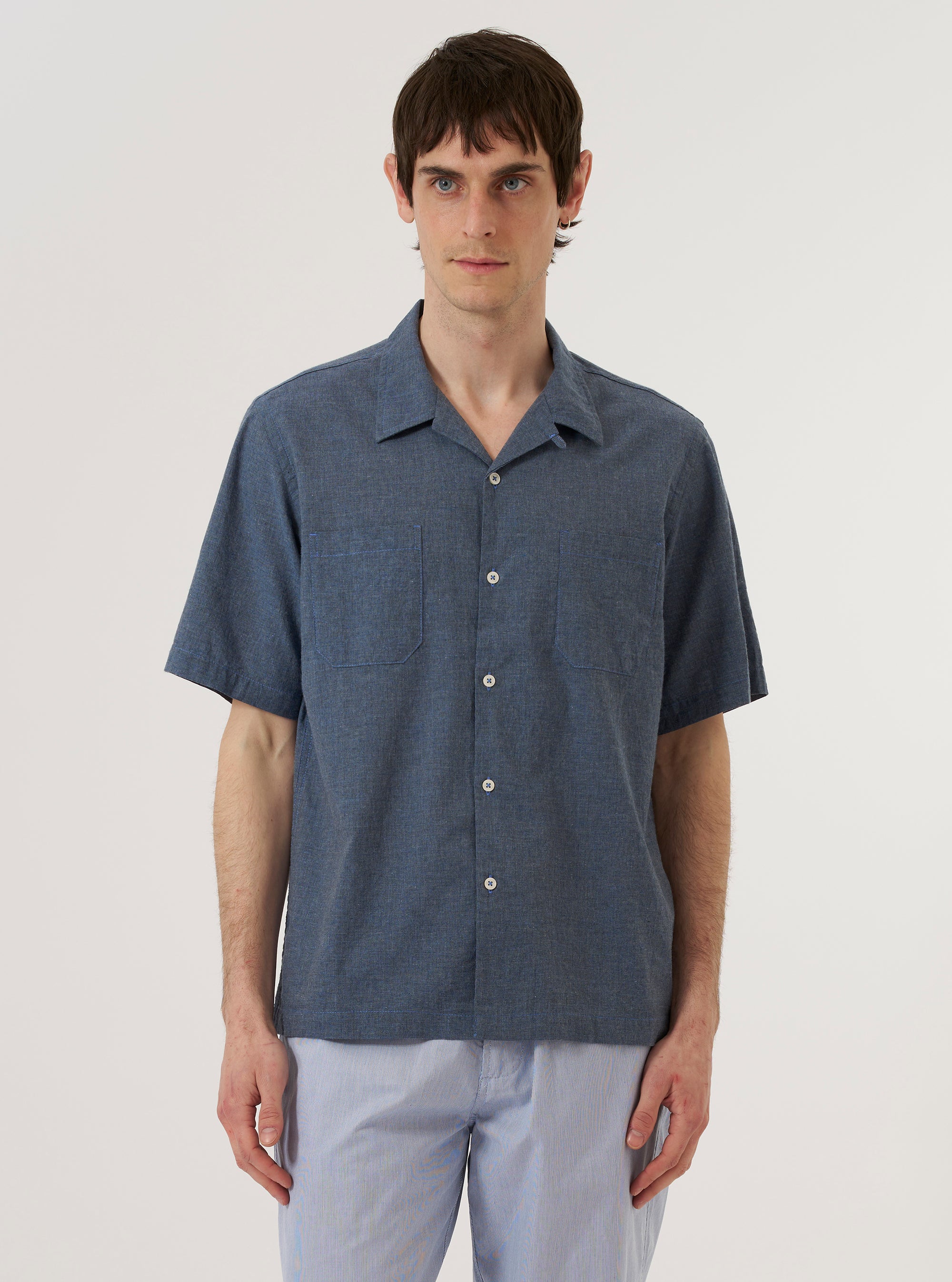 A model wears the Indigo Fine Organic Cotton Sal Road Shirt against a plain, neutral background. The shirt features a relaxed camp collar, a central button-down front, and a short-sleeved design. The garment is crafted from a fine indigo-dyed organic cotton fabric.
