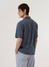 A model wears the Indigo Fine Organic Cotton Sal Road Shirt, shown from a rear three-quarter angle against a plain white background. The shirt features a regular fit with short sleeves, a straight hem, and a one-piece collar. The indigo blue organic cotton fabric has a visible marled texture and a lightweight, loose weave.