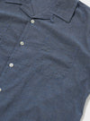 The Indigo Fine Organic Cotton Sal Road Shirt is shown from the back as a flat lay against a plain white background. The image highlights the garment's clean construction, featuring short sleeves, a straight hem, and the back of the camp collar. The organic cotton fabric is a deep indigo blue with a smooth, fine texture.