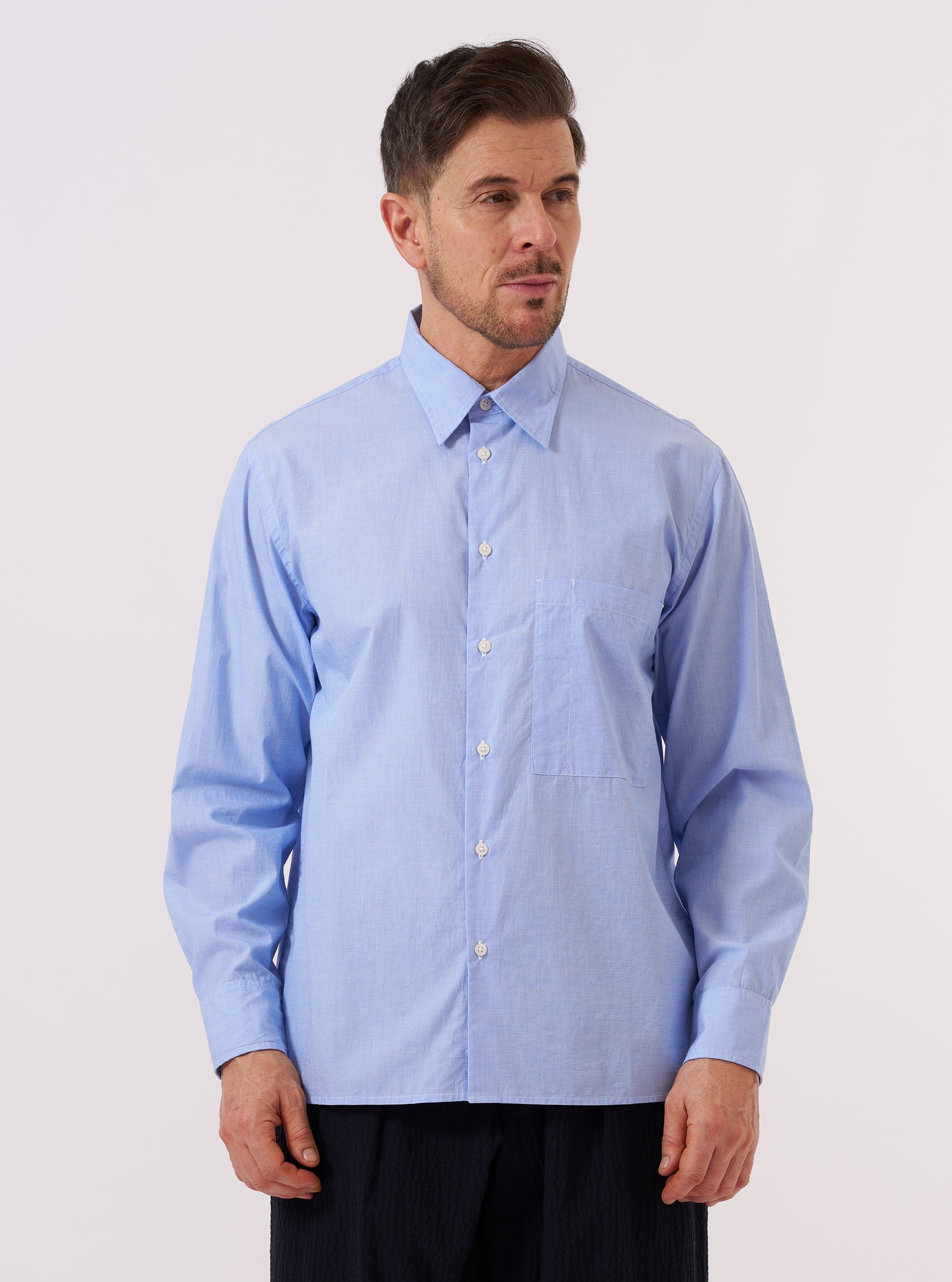 A model wears the Sky Fil A Fil Square Pocket Shirt against a plain, light-colored background. The shirt is a light sky blue with a fine end-on-end texture and features a prominent oversized square pocket on the left chest, a button-down front, and a classic collar.