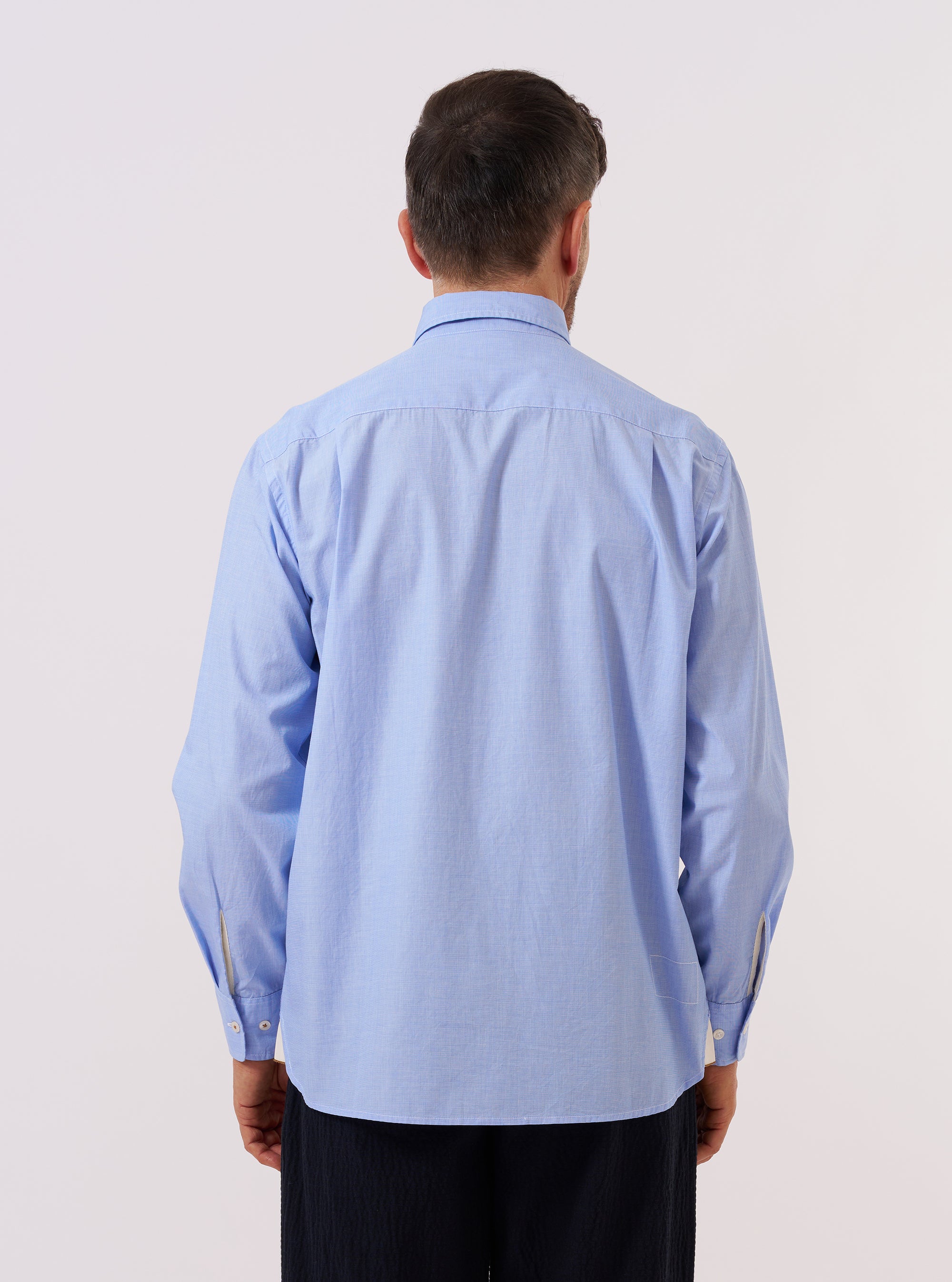 A model wears the Sky Fil A Fil Square Pocket Shirt against a neutral studio background. The shirt features a prominent square chest pocket on the left side and a classic button-down front with a pointed collar. The light blue end-on-end fabric weave is visible as the model poses to show the shirt's fit and length.