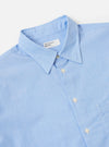 A model wears the Sky Fil A Fil Square Pocket Shirt against a plain, light-colored background. The light blue shirt is made from cotton with a subtle end-on-end weave and is shown from a slight angle to highlight its loose fit. Prominent features include a point collar, a full button-down front, and a large, oversized square patch pocket on the left chest. The long sleeves are finished with adjustable button cuffs, and the shirt has a visible curved hem.