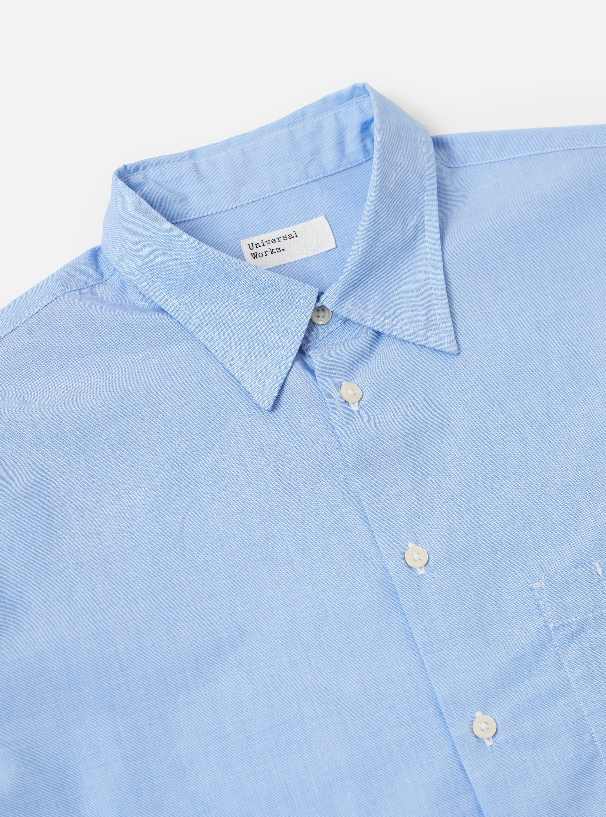 A model wears the Sky Fil A Fil Square Pocket Shirt against a plain, light-colored background. The light blue shirt is made from cotton with a subtle end-on-end weave and is shown from a slight angle to highlight its loose fit. Prominent features include a point collar, a full button-down front, and a large, oversized square patch pocket on the left chest. The long sleeves are finished with adjustable button cuffs, and the shirt has a visible curved hem.