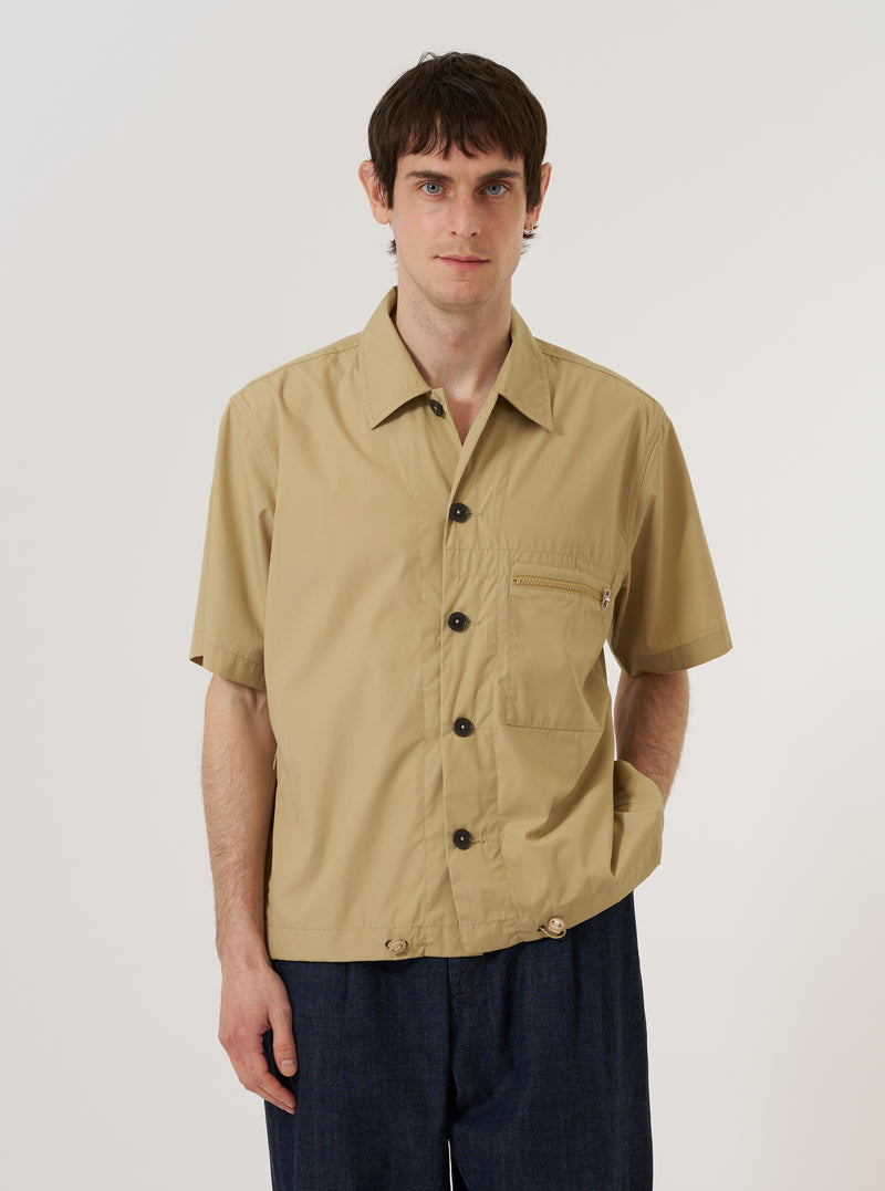 A model wears the Sand Recycled Polytech Z Tech Overshirt over a light-colored top, standing against a plain, light grey background. The utility-style overshirt features a classic collar, a central button-down front, and two large patch pockets at the waist. The sand-colored fabric has a smooth, technical finish.
