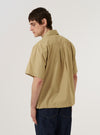 A model wears the Sand Recycled Polytech Z Tech Overshirt over a light-colored top, shown from a rear three-quarter angle against a plain white background. The image highlights the smooth, technical texture of the sand-colored fabric, the simple collar, and the straight hem. One large utility patch pocket is visible on the side of the garment.