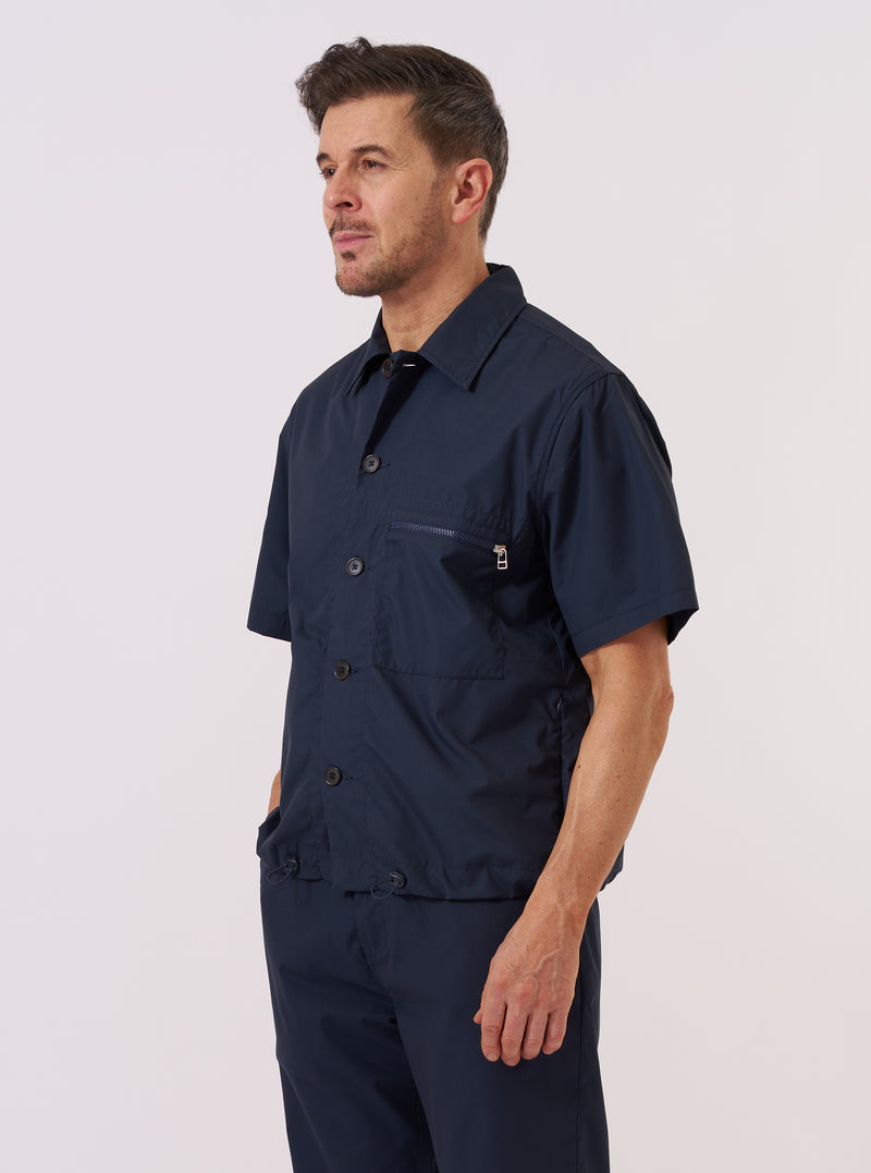 A model wears the Navy Recycled Polytech Z Tech Overshirt against a plain white background. The overshirt features a one-piece collar, a central button-down closure, and a prominent reinforced patch pocket on the left chest with a vertical zip. The dark navy fabric has a smooth, technical finish, and the garment is designed with a boxy, straight fit.