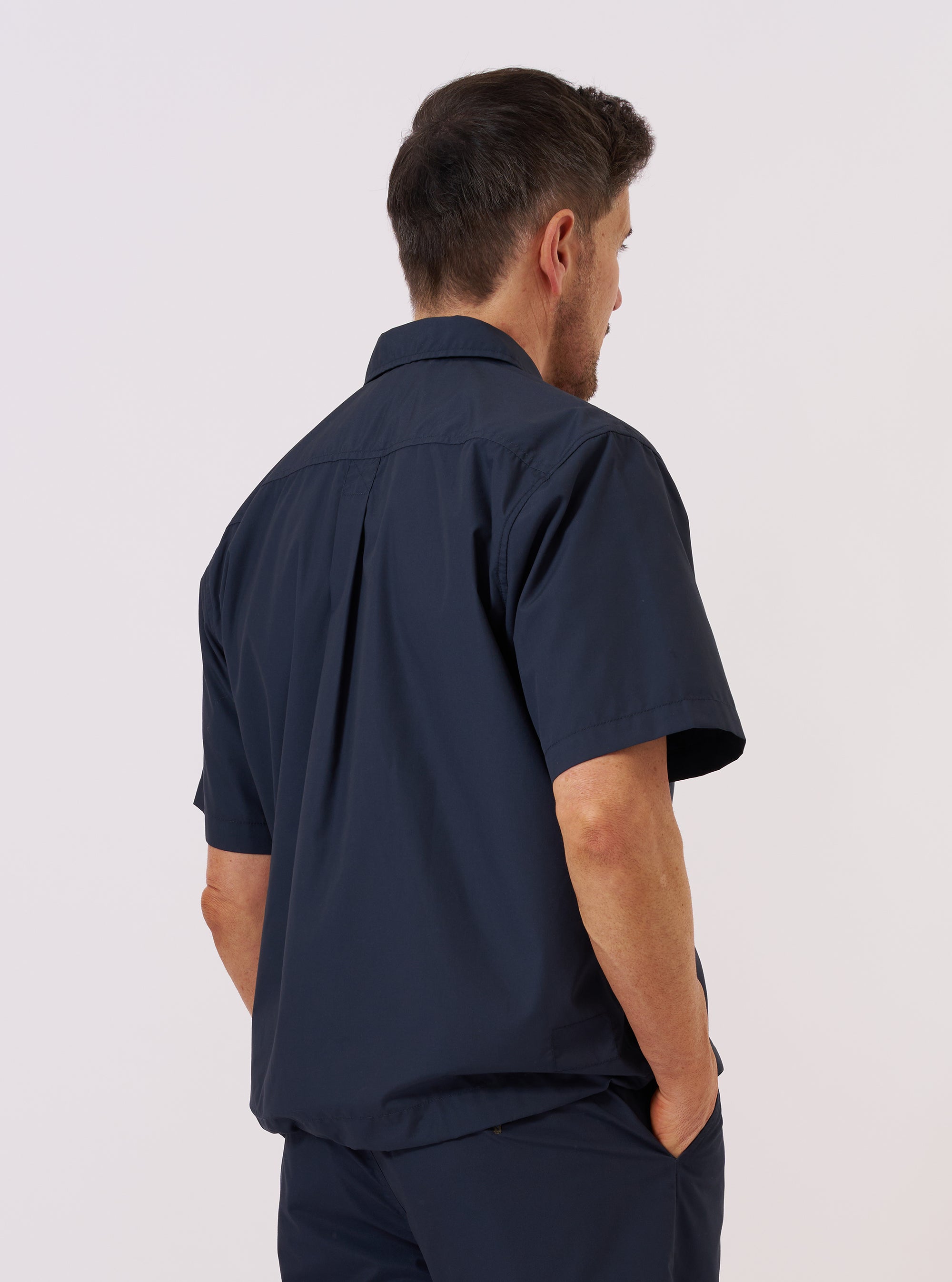 A model wears the Navy Recycled Polytech Z Tech Overshirt, shown from a rear three-quarter angle against a plain white background. The overshirt features a structured collar, long sleeves, and a clean, utility-style back construction with a straight hem. The dark navy fabric has a smooth, technical finish.