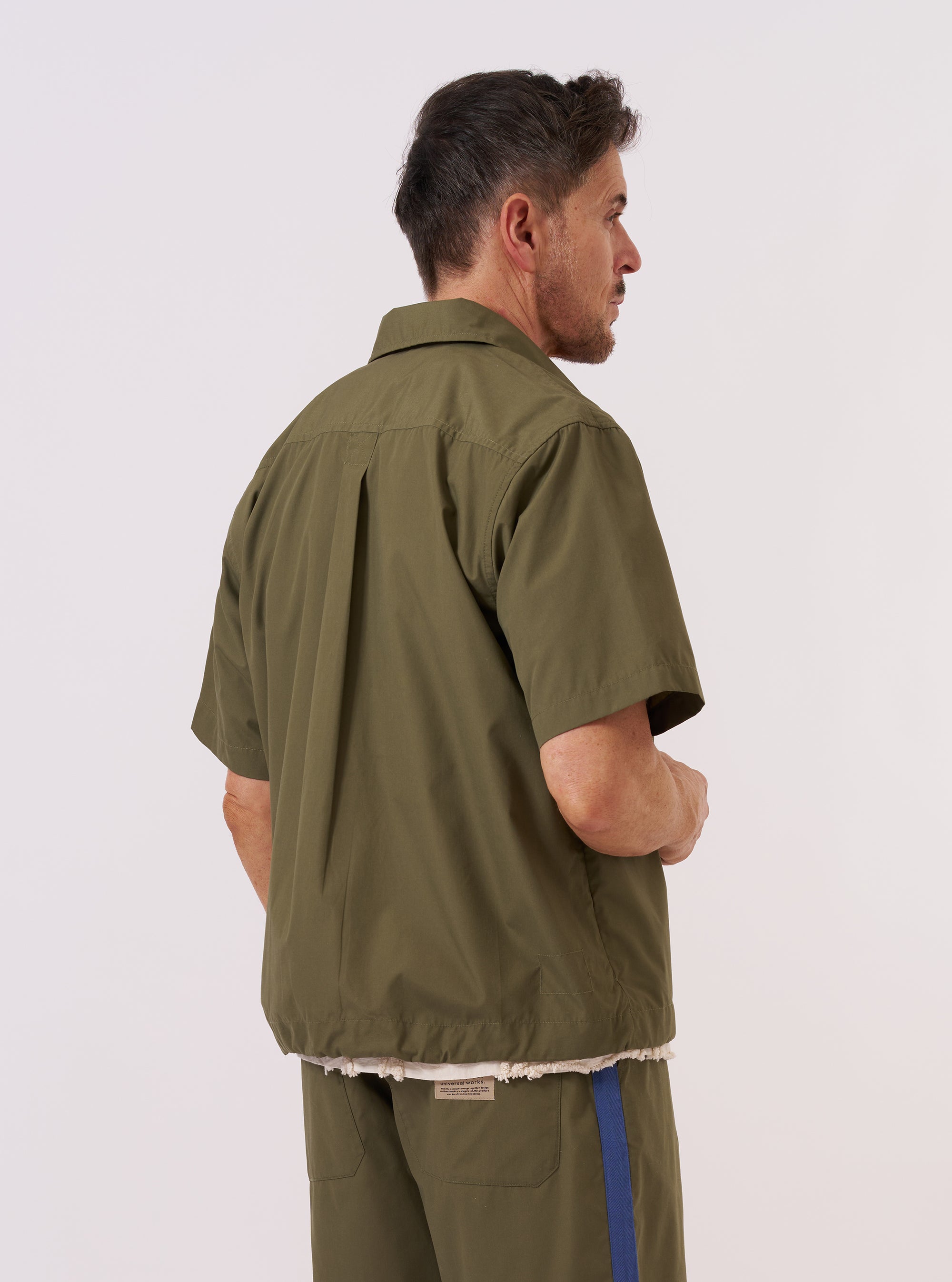A model wears the Olive Recycled Polytech Z Tech Overshirt over a light-colored top, shown from a rear three-quarter angle against a plain white background. The overshirt features a relaxed fit, a straight hem, and a simple collar. The smooth, olive green recycled polytech fabric is the prominent feature of the garment.