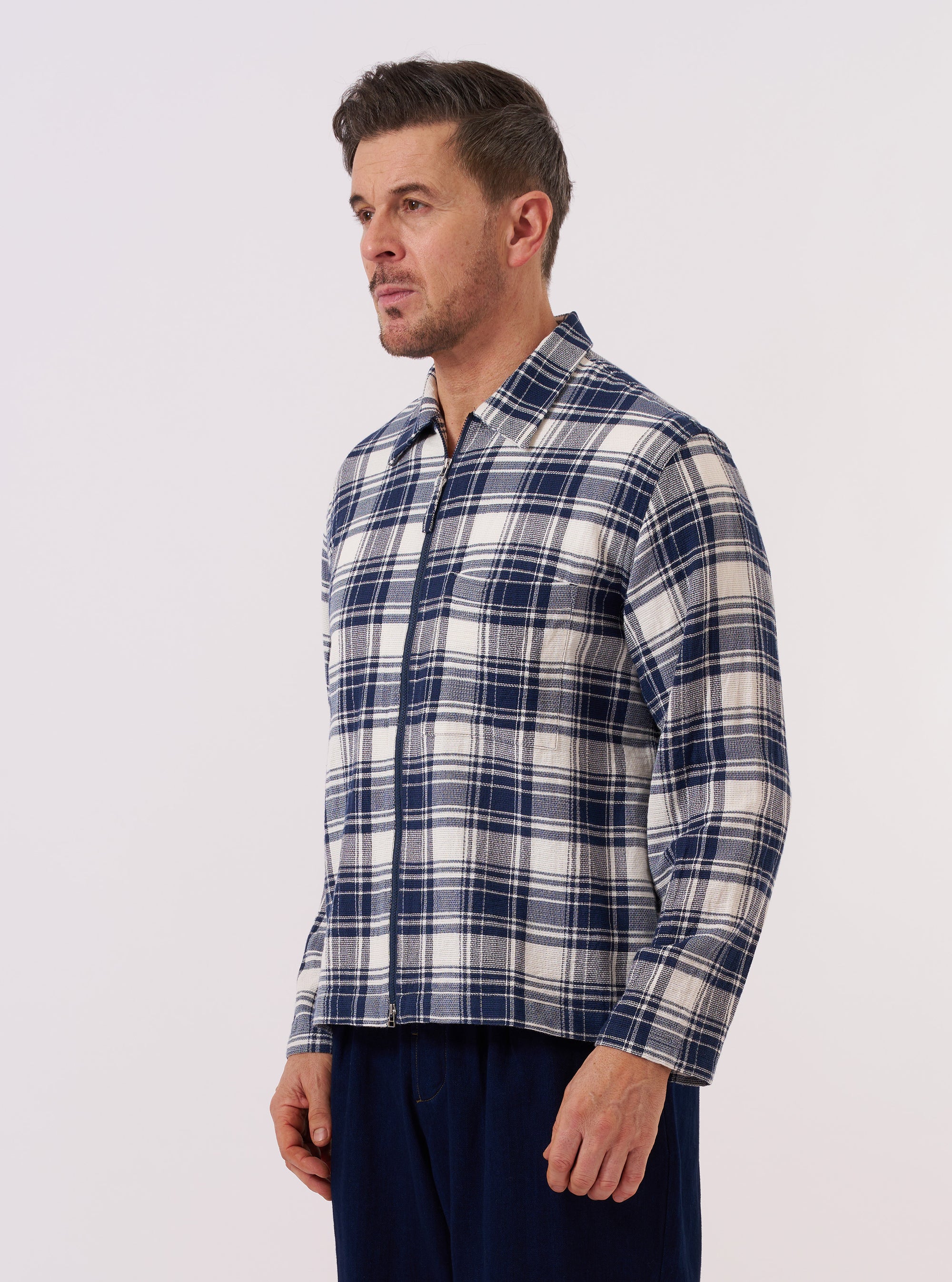 A model wears the Navy Texture Plaid Zip Shirt over a light-colored top, standing against a plain white background. The shirt features a full-length central zip closure, a classic pointed collar, and a textured navy blue plaid pattern. The garment is shown from the front, highlighting its relaxed fit and fabric detail.