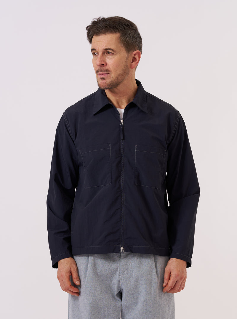 A model wears the Navy Seo Recycled Nylon Zip Too Shirt over a light-colored top against a plain, neutral background. The overshirt features a full-length front zipper, a classic collar, and two large patch pockets at the waist. The smooth, dark navy recycled nylon fabric has a subtle technical sheen.