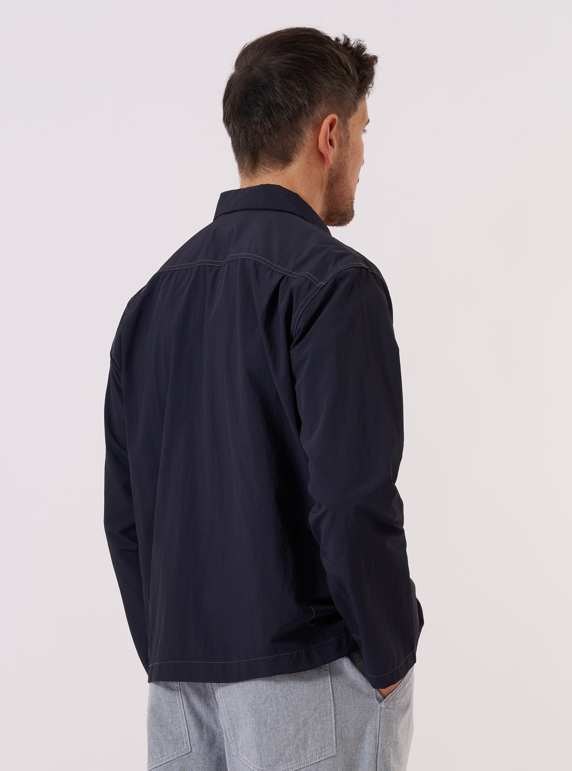 A model wears the Navy Seo Recycled Nylon Zip Too Shirt over a light-colored top, shown from a side-angle view against a plain, neutral studio background. The shirt features a smooth technical fabric made from recycled nylon, a pointed collar, and a full-length front zipper. The relaxed silhouette and deep navy color are prominent.