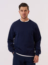 A model wears the Navy Brush Back Sweat Billy Sweat over a light-colored shirt, standing against a plain, neutral studio background. The sweatshirt features a classic crew neckline, ribbed cuffs, and a ribbed hem. The fabric is a smooth, dark navy cotton with a visible soft texture.