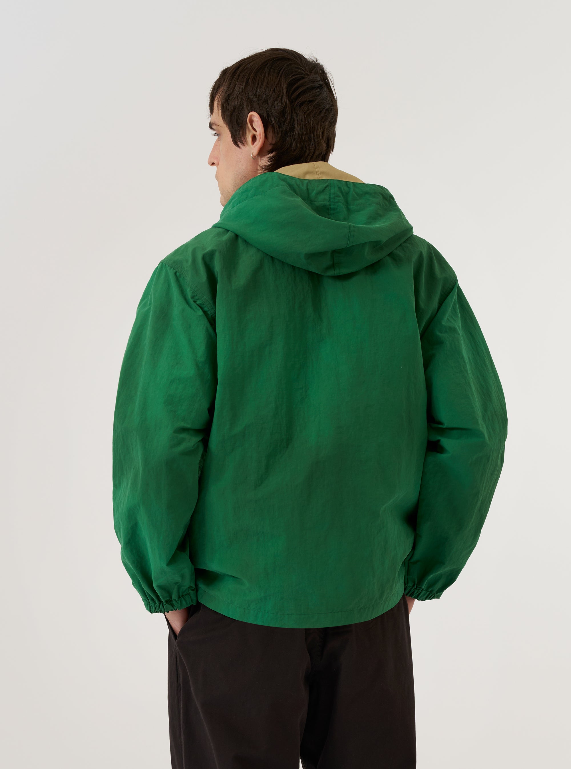A model wears the Green Recycled Splash Nylon Longsands Jacket against a plain white background. The image shows the back of the garment, highlighting the integrated hood, long sleeves, and the smooth, lightweight texture of the green recycled nylon fabric. The jacket features a relaxed fit with a straight hem. No mannequin is present in the image.