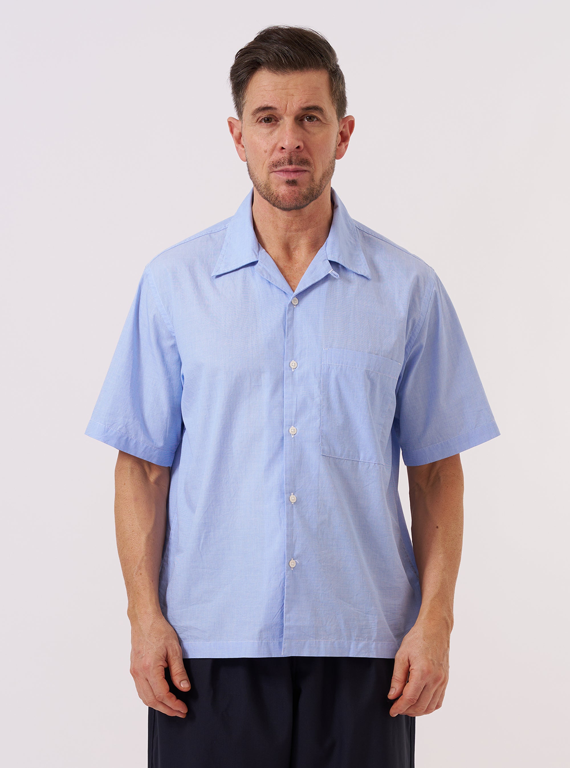 A model wears the Sky Fil A Fil Camp Shirt against a plain, neutral background. The shirt features a relaxed camp collar, a central button-down front, and a single patch pocket on the left chest. The garment is made from a light sky blue cotton fil-a-fil fabric. The model is shown in a front-facing view, wearing the shirt as a top layer.