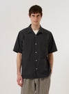A model wears the Black Tech Cotton S/S Dwight Overshirt against a plain white background. The short-sleeved overshirt features a camp collar, a button-up front, and two large utility-style patch pockets on the chest. The model wears the garment unbuttoned over a white t-shirt, showcasing its relaxed fit and the smooth, technical texture of the black cotton fabric.