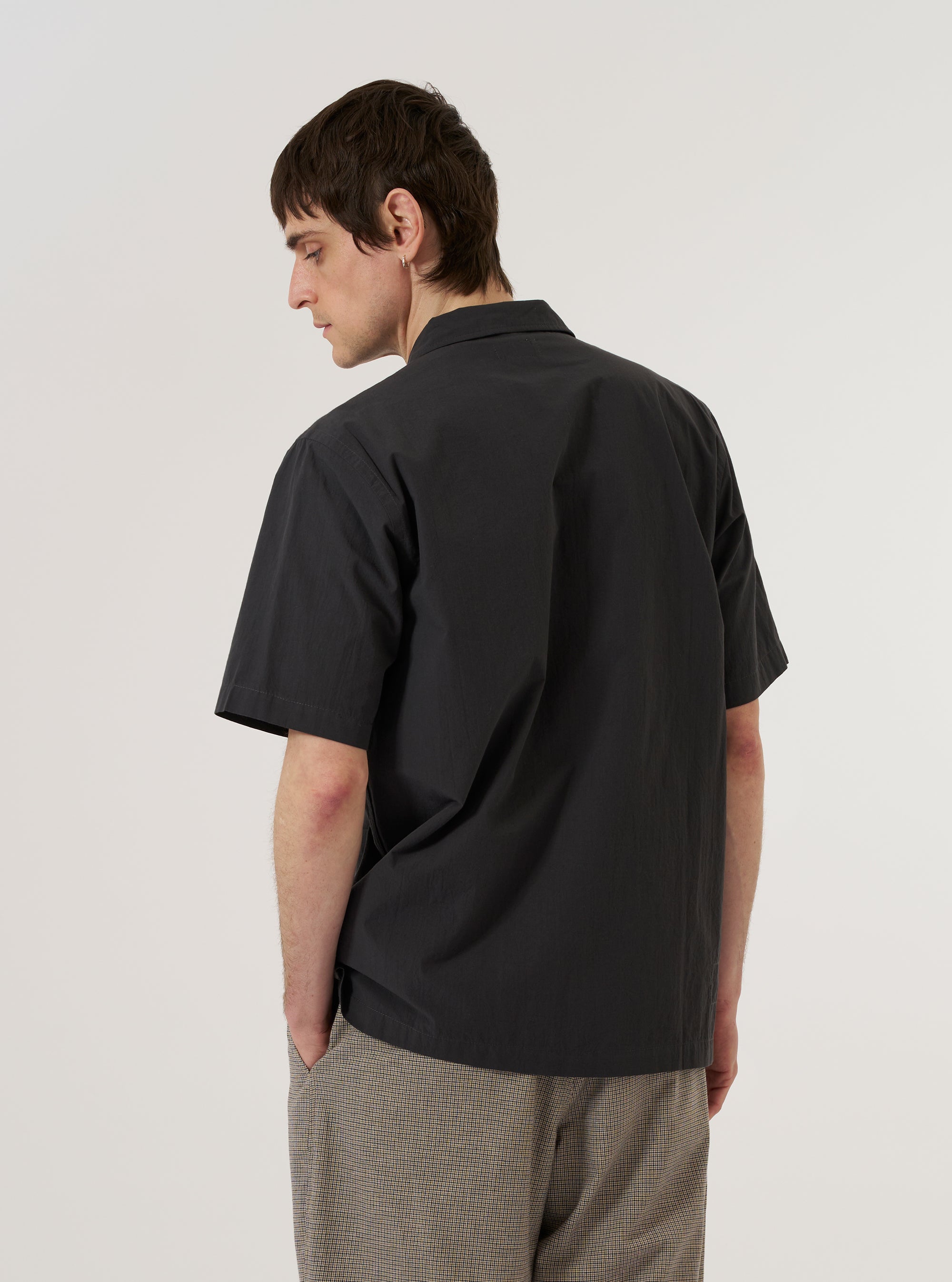 A model wears the Black Tech Cotton S/S Dwight Overshirt over a light-colored top, shown from a rear three-quarter angle against a plain white background. The short-sleeve overshirt features a structured camp collar and a straight hem. The black tech cotton fabric has a smooth, technical appearance.