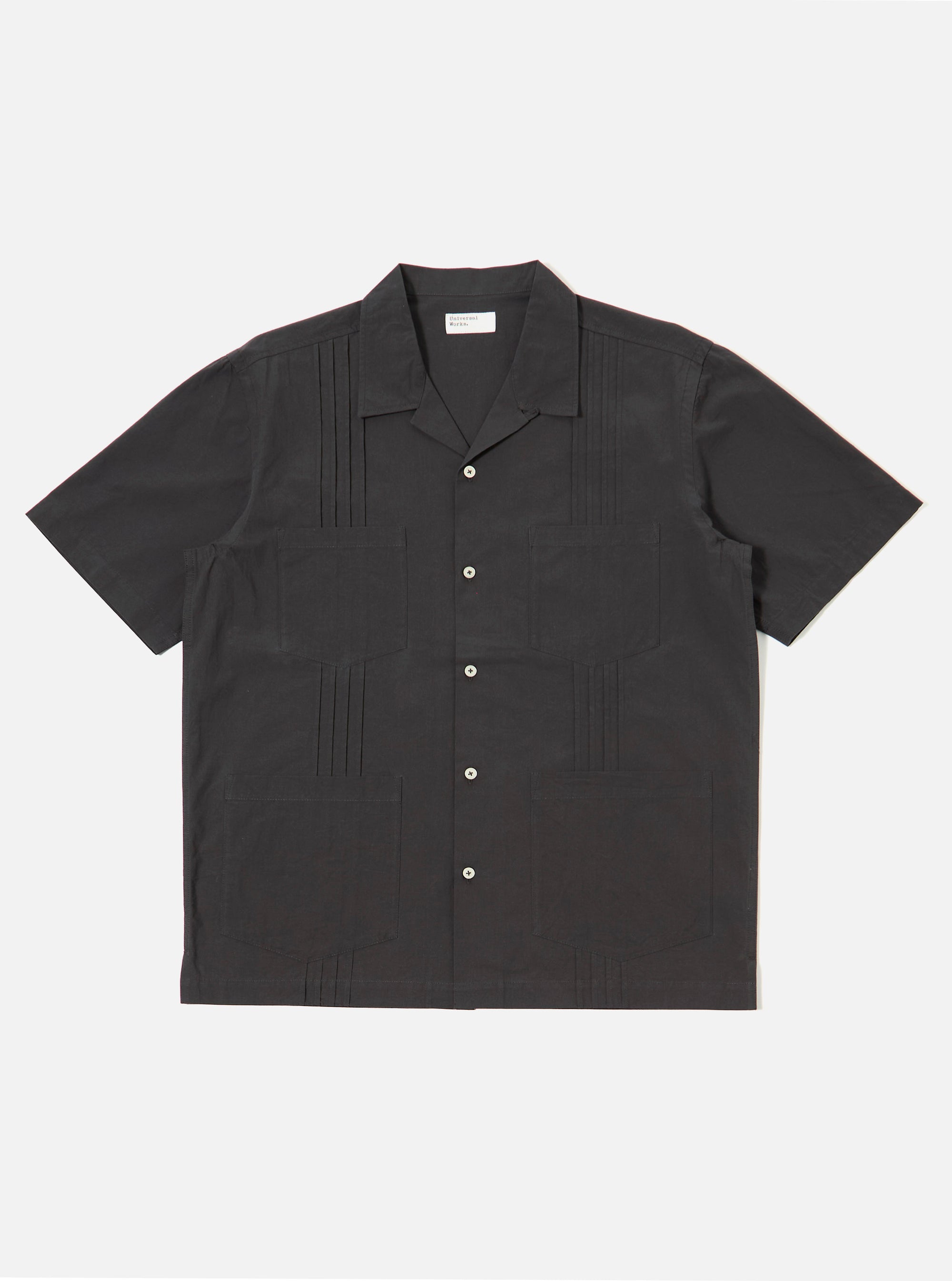 The Black Tech Cotton S/S Dwight Overshirt is shown as a flat lay against a white background. The short-sleeved overshirt features a straight hem, a button-down front with a camp collar, and four front patch pockets. Two rows of vertical sewn pleats run down the front on either side of the buttons. The fabric is a smooth black tech cotton.