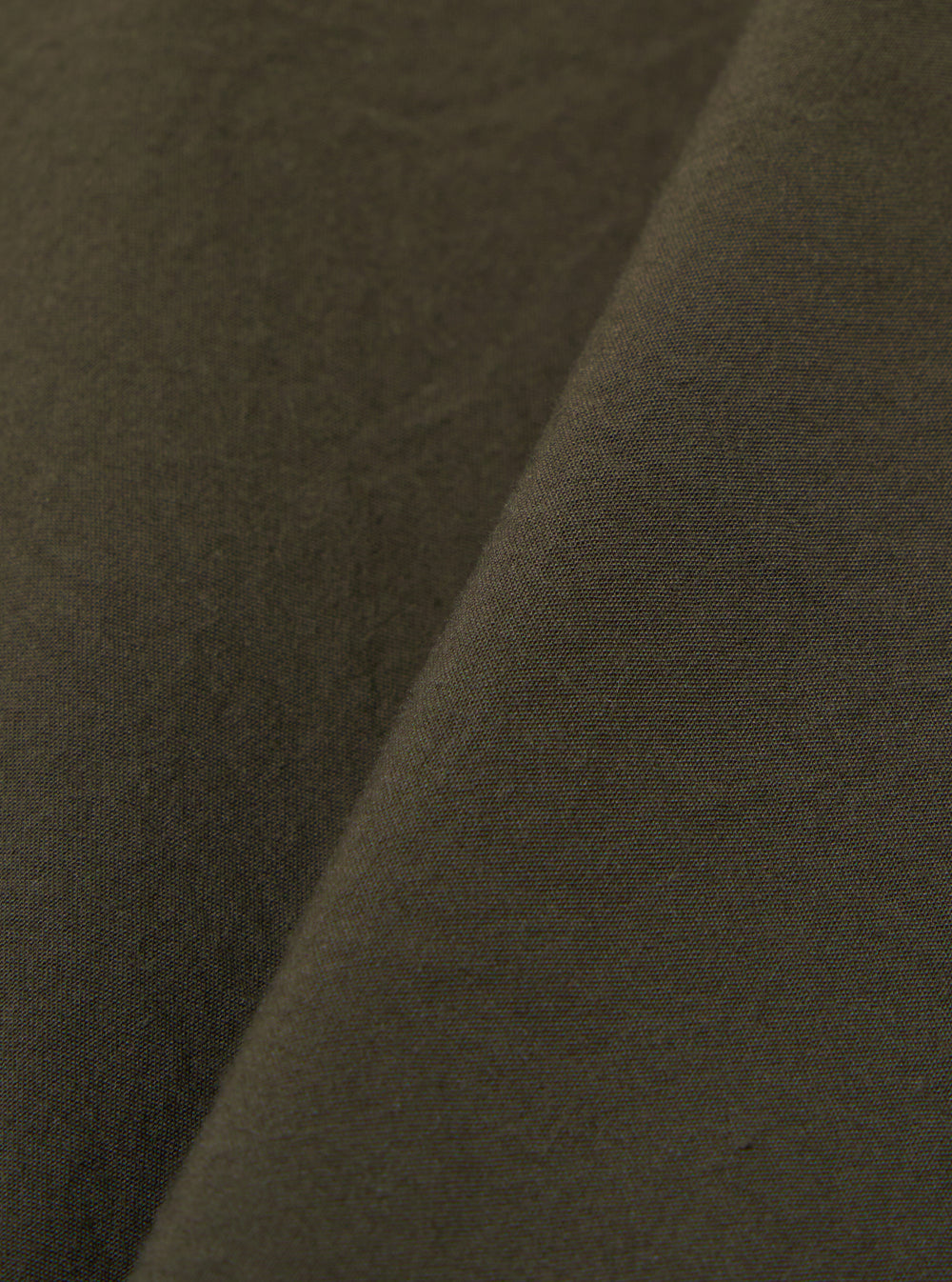 A close-up fabric swatch of the Olive Tech Cotton S/S Dwight Overshirt highlights the olive green color and the smooth texture of the tech cotton fabric. The material fills the frame, providing a detailed view of the garment's construction.