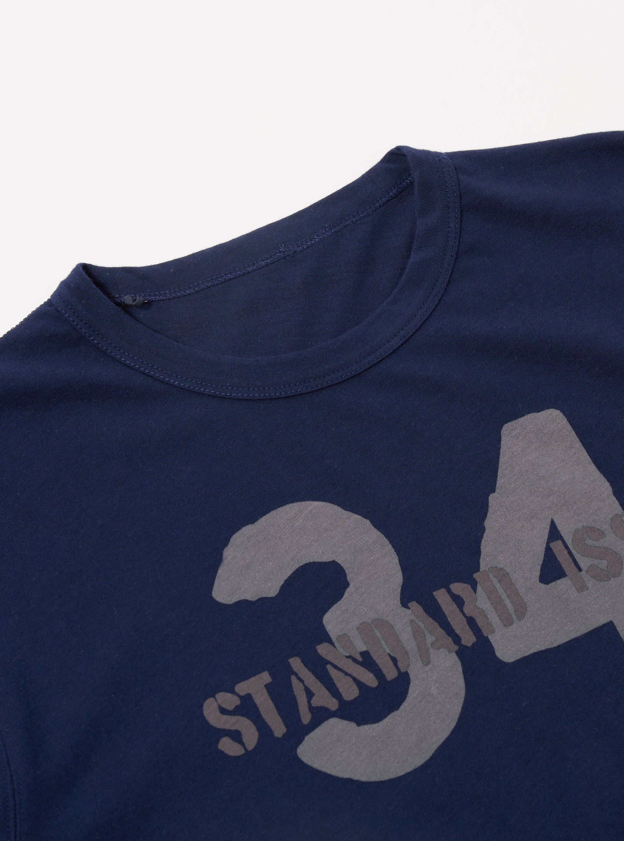 The Navy Single Jersey 34 Printed Core Tee is shown in a close-up detail shot against a plain white background. The image focuses on the chest area of the navy blue cotton garment, highlighting the white graphic print and the fine texture of the single jersey fabric. The ribbed crew neck is partially visible at the top.