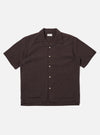 The Brown Diamond Dobby Deck Shirt is shown as a flat lay against a plain white background. The shirt features a one-piece collar, a central button-down front, and two large patch pockets at the waist. The brown cotton fabric has a distinct diamond dobby texture and the garment is designed with short, loose sleeves and a straight hem.