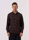A model wears the Brown Diamond Dobby Lucky Shirt over a light-colored t-shirt, standing against a plain, neutral background. The shirt features a short-sleeve design, a classic collar, and a central button closure. The brown cotton fabric is characterized by a visible, textured diamond dobby geometric pattern.