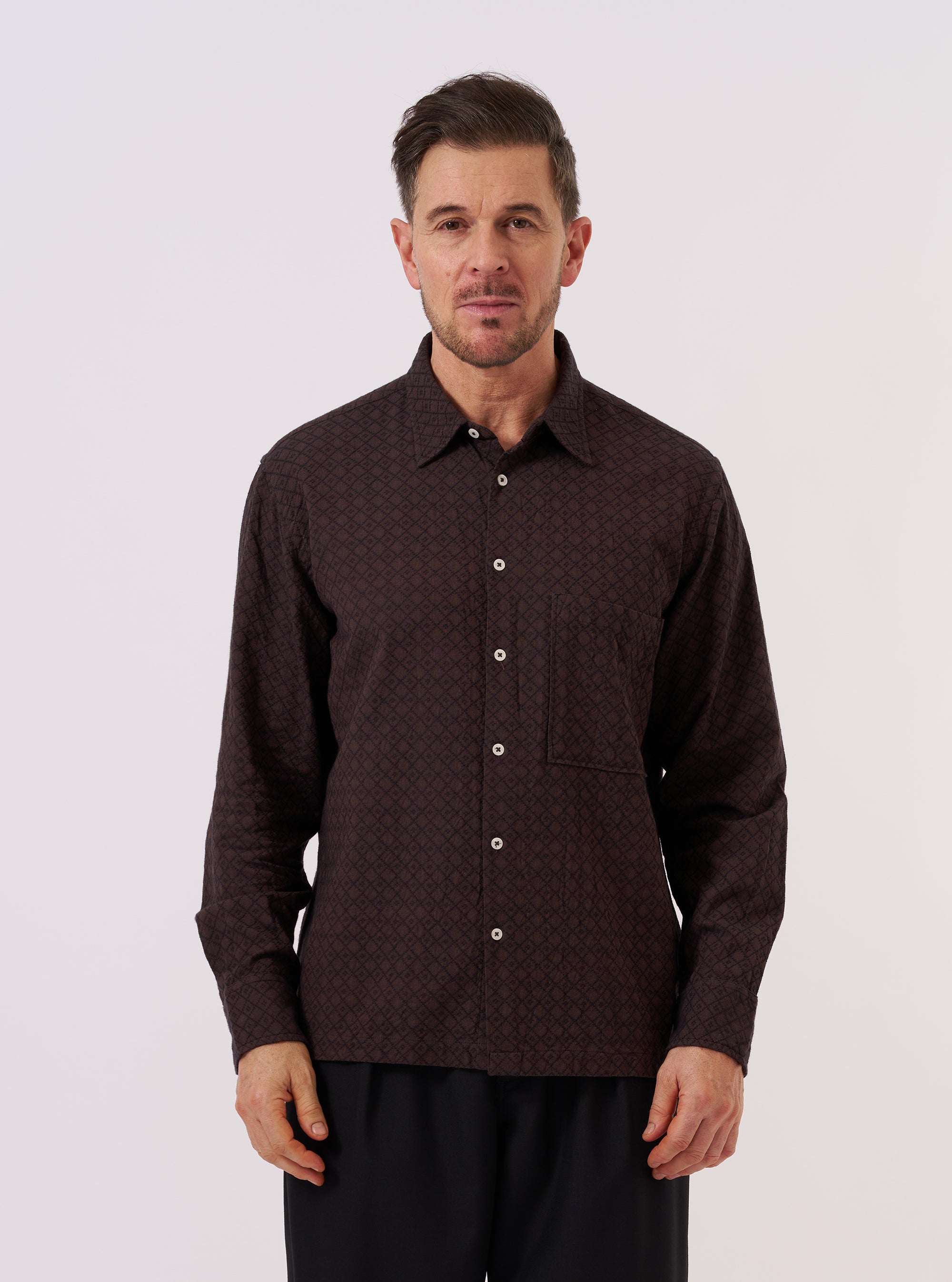 A model wears the Brown Diamond Dobby Lucky Shirt over a light-colored t-shirt, standing against a plain, neutral background. The shirt features a short-sleeve design, a classic collar, and a central button closure. The brown cotton fabric is characterized by a visible, textured diamond dobby geometric pattern.