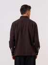A model wears the Brown Diamond Dobby Lucky Shirt, shown from a rear three-quarter angle against a plain, neutral studio background. The shirt features a relaxed silhouette, buttoned cuffs, and a straight hem. The brown cotton fabric is characterized by a distinct and consistent textured diamond dobby pattern throughout.