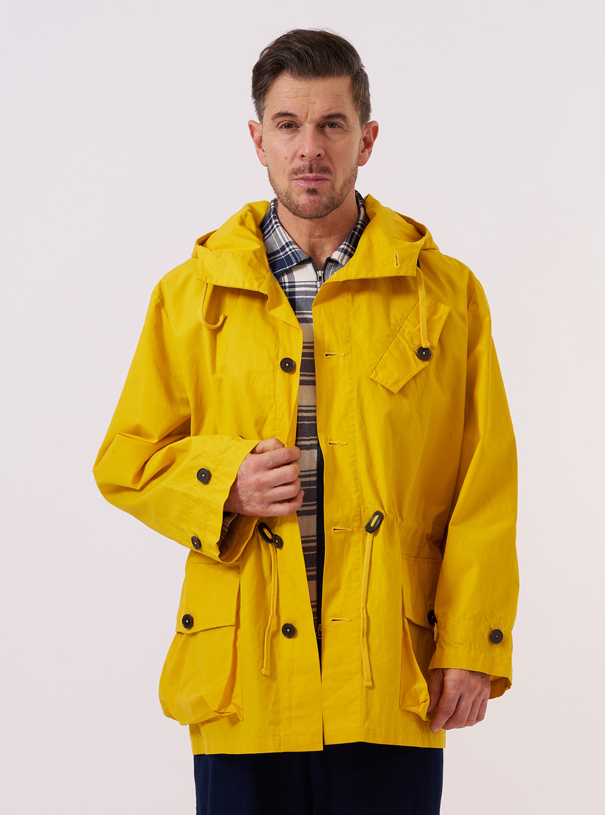 A model wears the Yellow British Wax Cotton Boston Parka against a plain white background. The oversized jacket features a vibrant yellow ochre color, a large drawstring hood, and two prominent 3D bellows pockets at the waist. It has a central button-down storm flap over a zipper and long sleeves with adjustable cuffs. The model is shown from the front, highlighting the parka's sculptural proportions and lightweight waxed cotton texture.