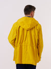 A model is wearing the Yellow British Wax Cotton Boston Parka against a plain white background, shown from the back. The parka is a vibrant yellow color and features an oversized silhouette with a large hood, a straight hem, and an inverted box pleat at the center back. The model is facing away from the camera, highlighting the garment's generous proportions and clean construction. No mannequin is present in the image.