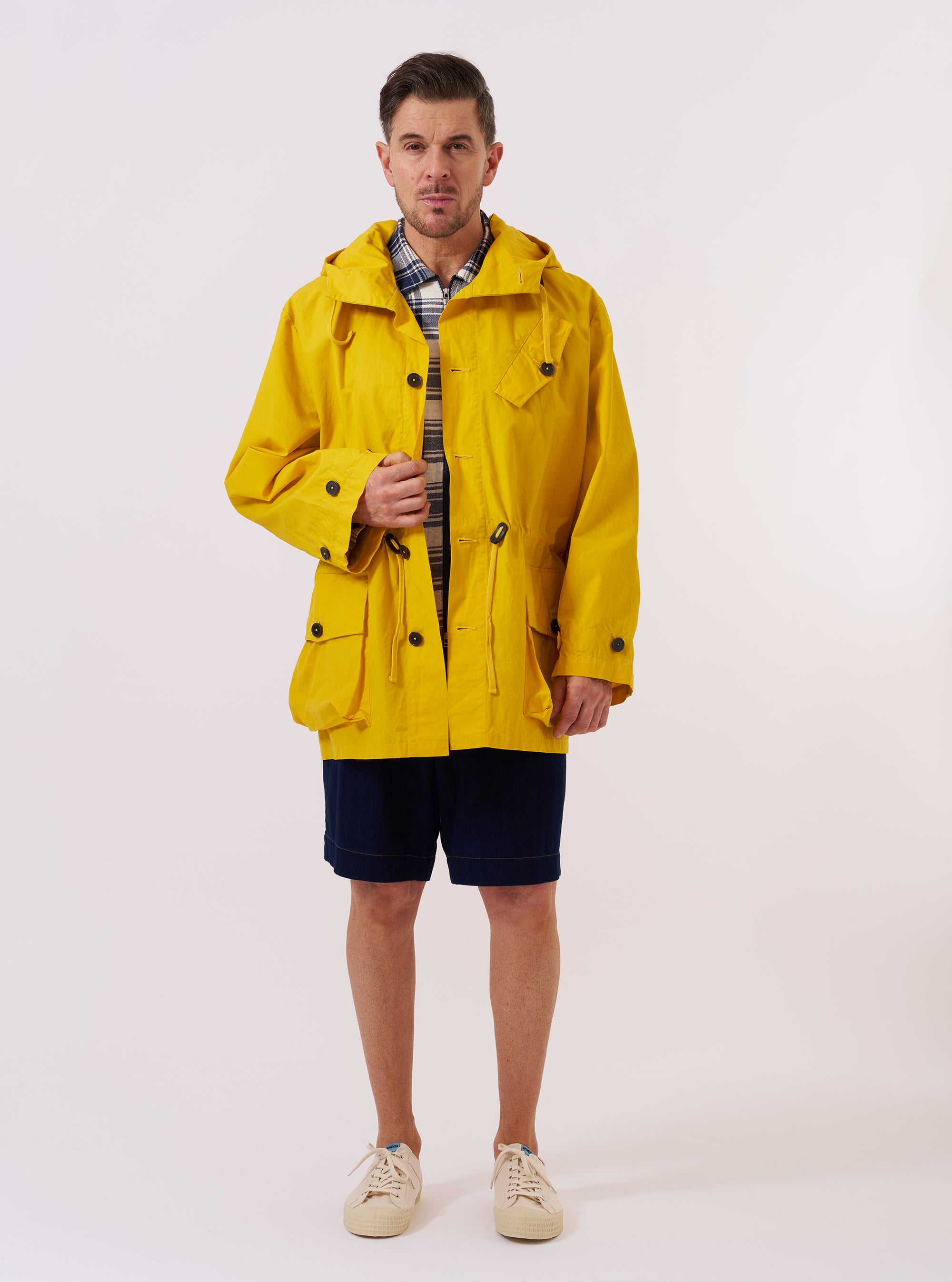 The Yellow British Wax Cotton Boston Parka is displayed as a flat lay against a plain white background. The parka features a high-neck hood, a central front fastening with a storm flap, and two spacious flap pockets at the hips. The garment is made from a bright yellow waxed cotton fabric that has a smooth, functional appearance.