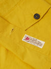 The Yellow British Wax Cotton Boston Parka is shown as a flat lay against a plain white background. The parka features an oversized silhouette with a large hood, a central button-down storm flap, one slanted chest pocket, and two prominent 3D patch pockets at the hip. The garment is constructed from a matte yellow waxed cotton fabric with visible tonal stitching and adjustable button cuffs.
