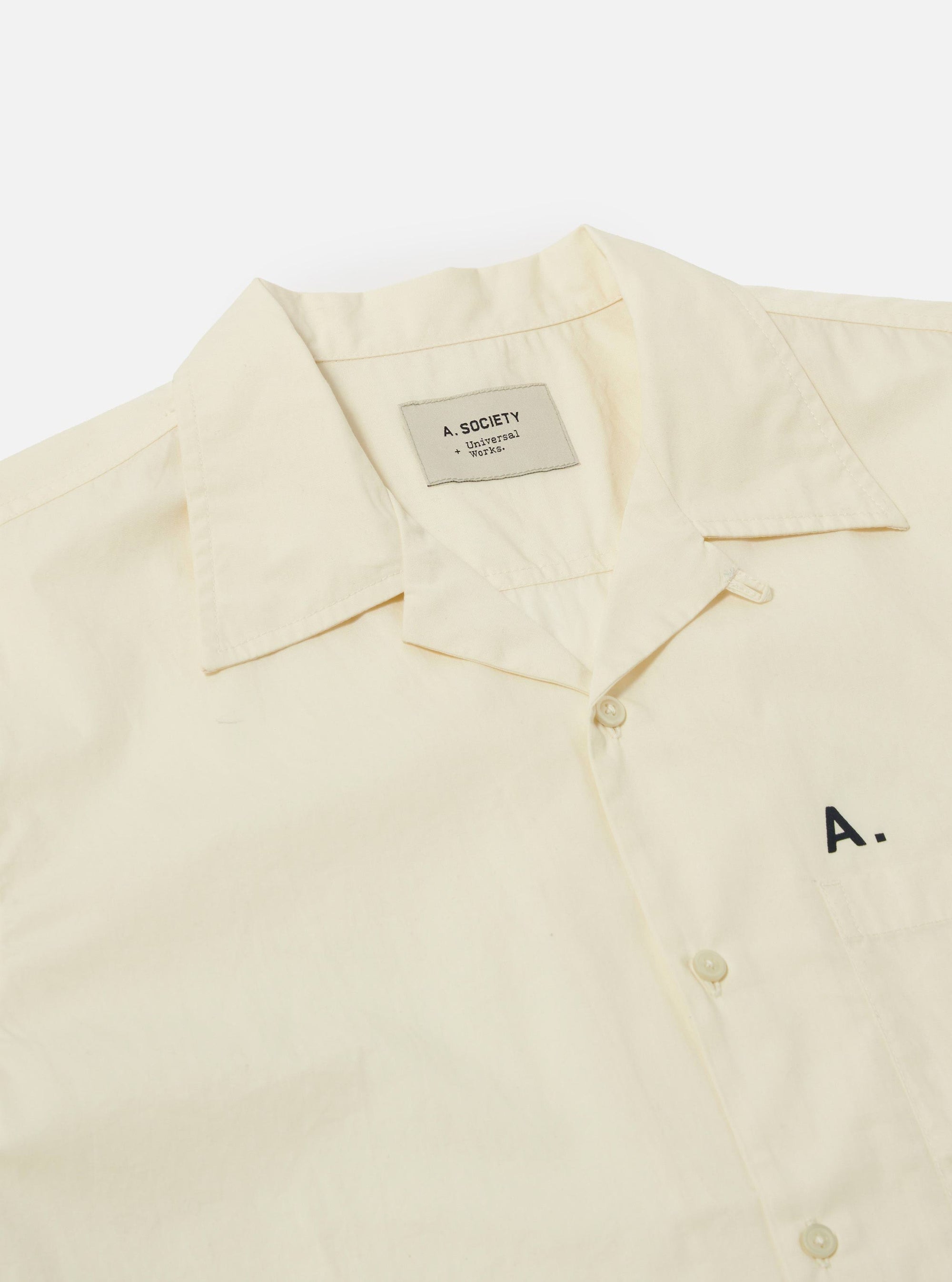Close-up of the A. SOCIETY x Universal Works Ecru Broadcloth Camp Shirt on a white background. The shirt is ecru-colored with a camp collar and button-up front. 'A.' is embroidered on the pocket.
