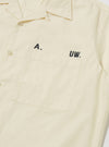 Close-up of the A. SOCIETY x Universal Works Ecru Broadcloth Camp Shirt. The ecru shirt features a chest pocket and black lettering. Shot on a plain white background.