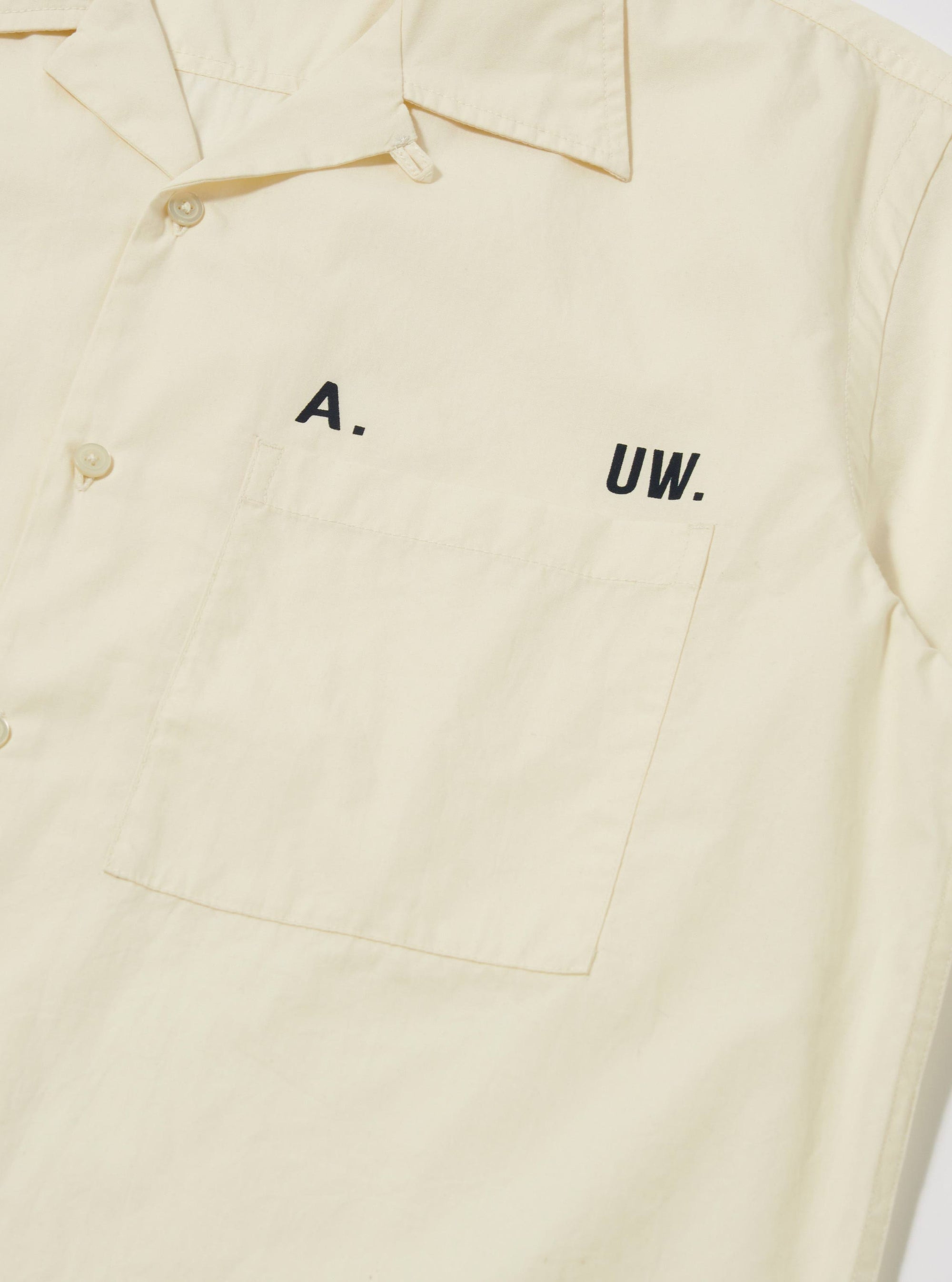 Close-up of the A. SOCIETY x Universal Works Ecru Broadcloth Camp Shirt. The ecru shirt features a chest pocket and black lettering. Shot on a plain white background.