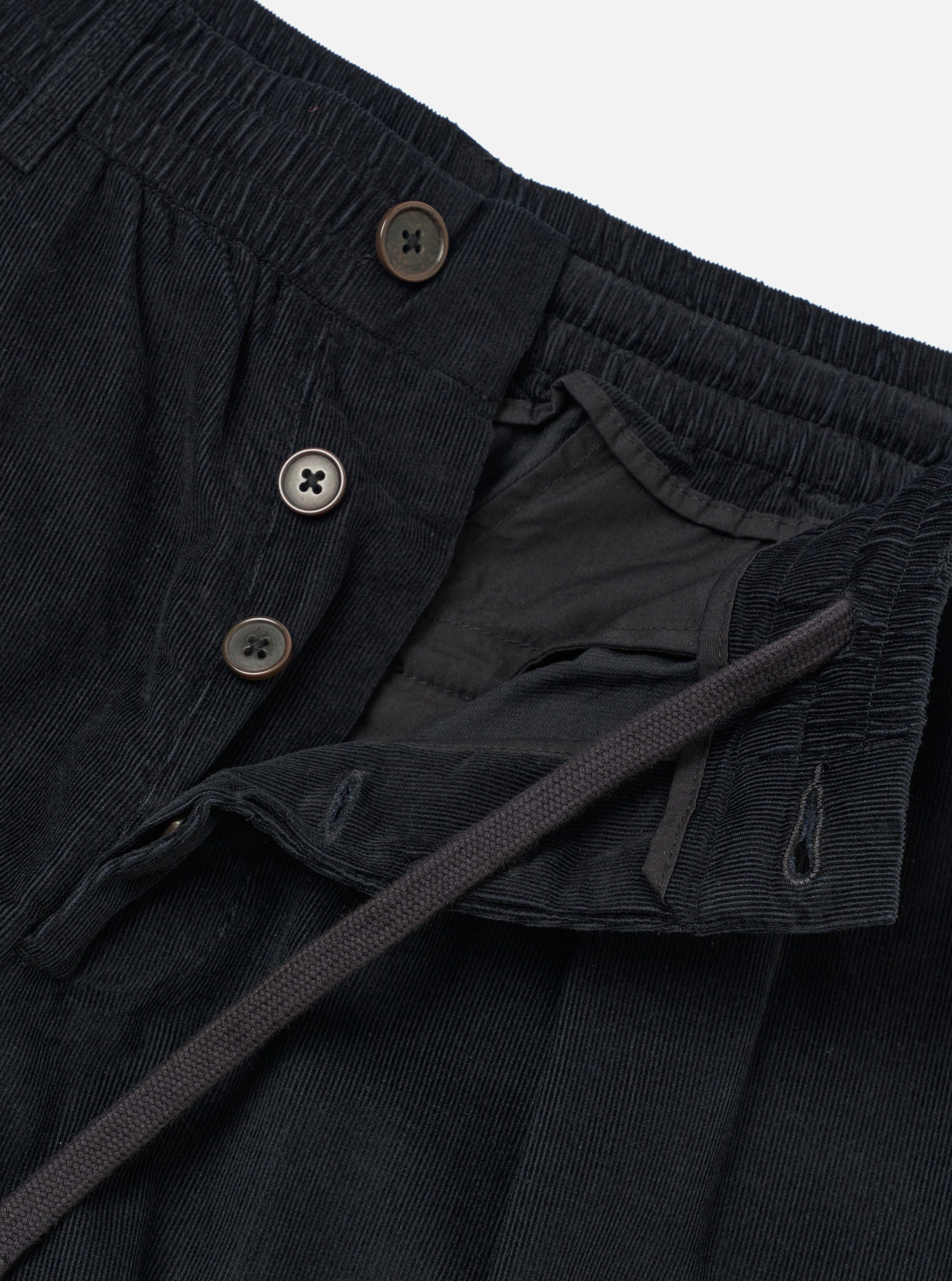The Black Organic Fine Cord Oxford Pant is shown as a flat lay against a plain white background. The image focuses on the rear of the trousers, highlighting the elasticated waistband, a buttoned flap pocket on the left, and a buttoned jetted pocket on the right. The fine-ribbed texture of the black organic cotton corduroy is visible throughout the garment.