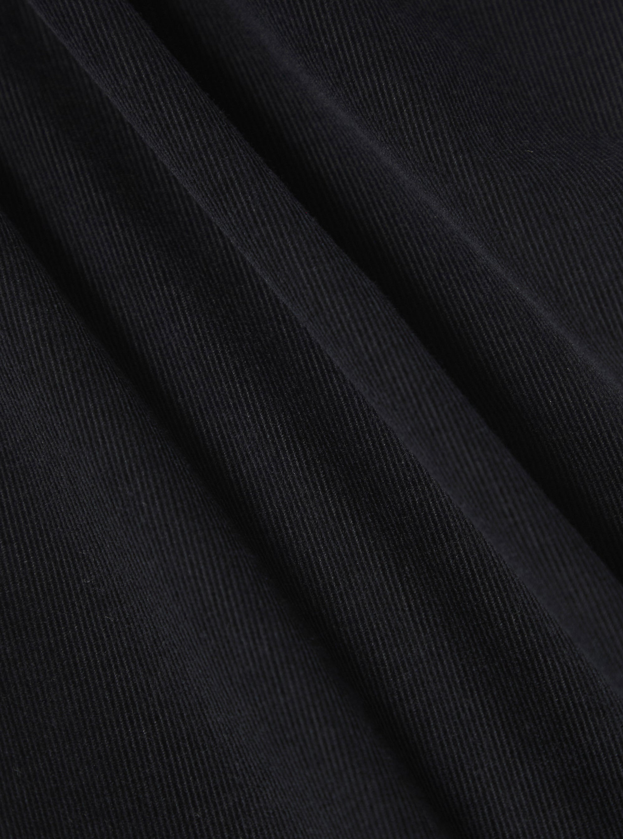 A close-up fabric swatch of the Black Organic Fine Cord Oxford Pant displays the black color and the fine, ribbed texture of the organic cotton corduroy material. The fabric fills the frame, highlighting its detailed construction.