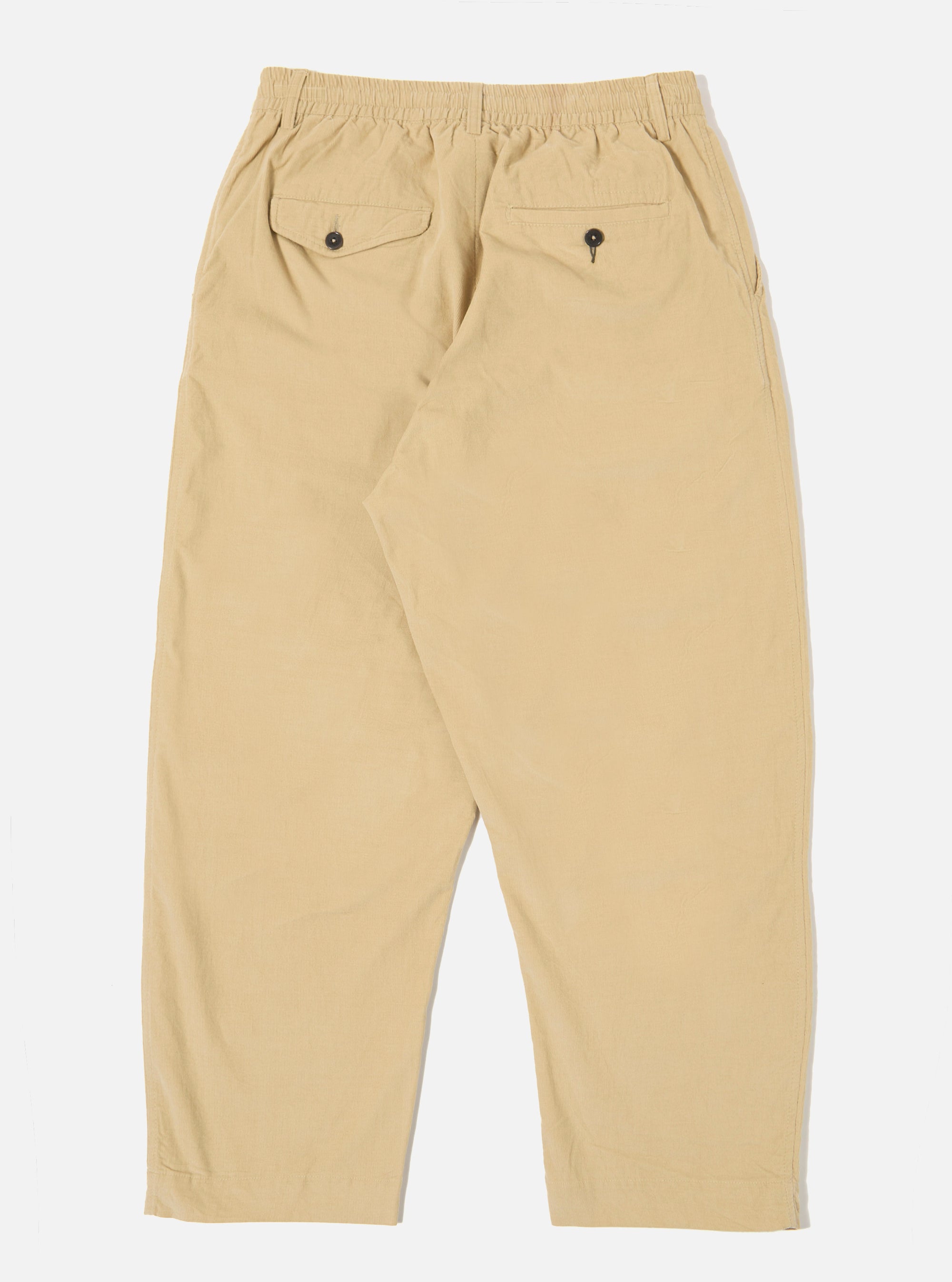 The Sand Organic Fine Cord Oxford Pant is shown from the back as a flat lay against a plain white background. The image highlights the two rear pockets, including one with a buttoned flap and one jetted pocket, as well as the elasticated waistband. The sand-colored organic cotton fabric features a visible fine corduroy texture.