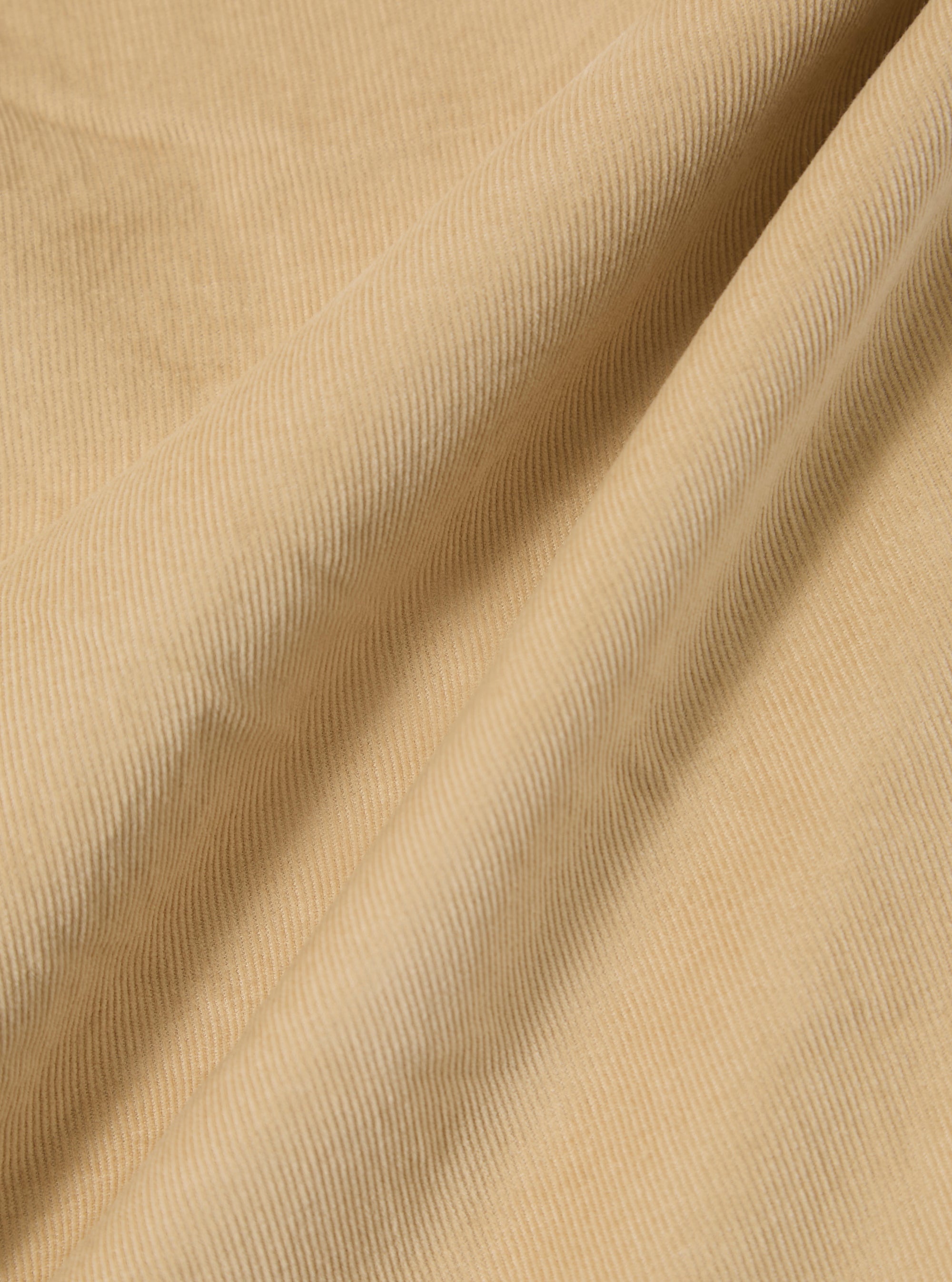 A close-up swatch of the Sand Organic Fine Cord Oxford Pant highlights the fine, vertical 21 wale texture of the organic cotton corduroy. The fabric is a solid, light sand color.