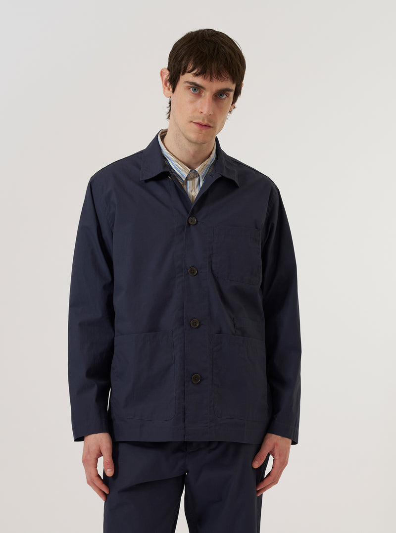 A model wears the Navy Peached Poplin Bakers Overshirt over a light-colored top, standing against a plain white background. The overshirt features a classic one-piece collar, a button-down front, and four front patch pockets, including one asymmetric chest pocket and two large lower pockets. The dark navy cotton fabric has a soft, peached finish.