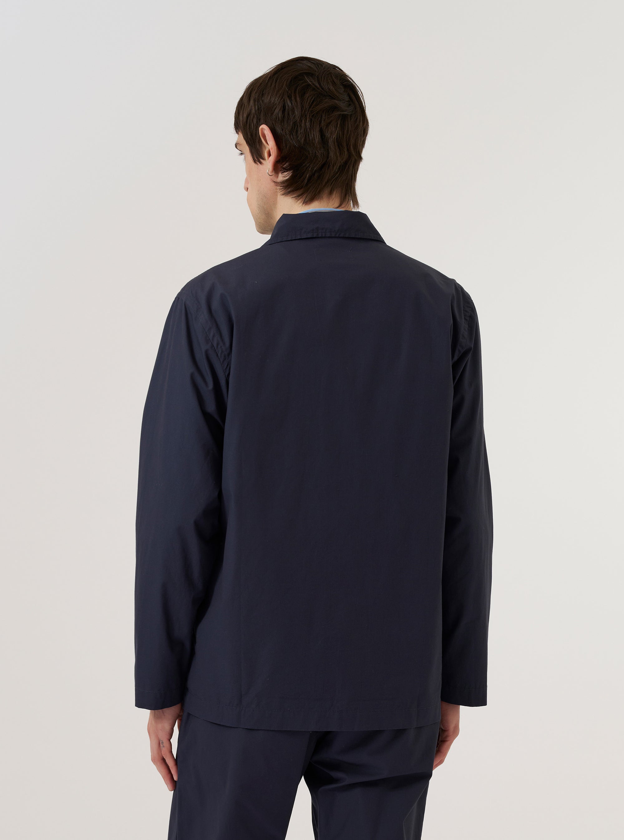 A model wears the Navy Peached Poplin Bakers Overshirt over a light-colored top, shown from a rear three-quarter angle against a plain white background. The overshirt features a relaxed fit, a straight hem, and a simple one-piece collar. The dark navy cotton poplin fabric has a smooth, peached finish.