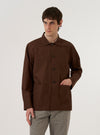 A model wears the Brown Peached Poplin Bakers Overshirt over a light-colored shirt, standing against a plain white background. The overshirt features a classic collar, a five-button front closure, and multiple front patch pockets, including one at the chest and two at the waist. The cotton fabric has a smooth, matte peached finish in a rich brown color.