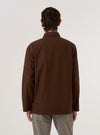 A model wears the Brown Peached Poplin Bakers Overshirt while standing against a plain white background. The image shows the garment from the back, highlighting its straight hem, relaxed boxy fit, and the smooth matte texture of the brown cotton poplin fabric. The model's hands are placed in the front pockets, partially visible from the side.