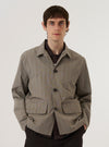 A model wears the Grey Brown Wales Glen Check Franck Jacket over a light-colored top, standing against a plain white background. The jacket features a shorter cut, a one-piece flat collar worn open, and two large front flap pockets. The fabric displays a detailed grey and brown glen check pattern. The model is shown from the front, highlighting the garment's workwear-inspired silhouette and button-down closure.