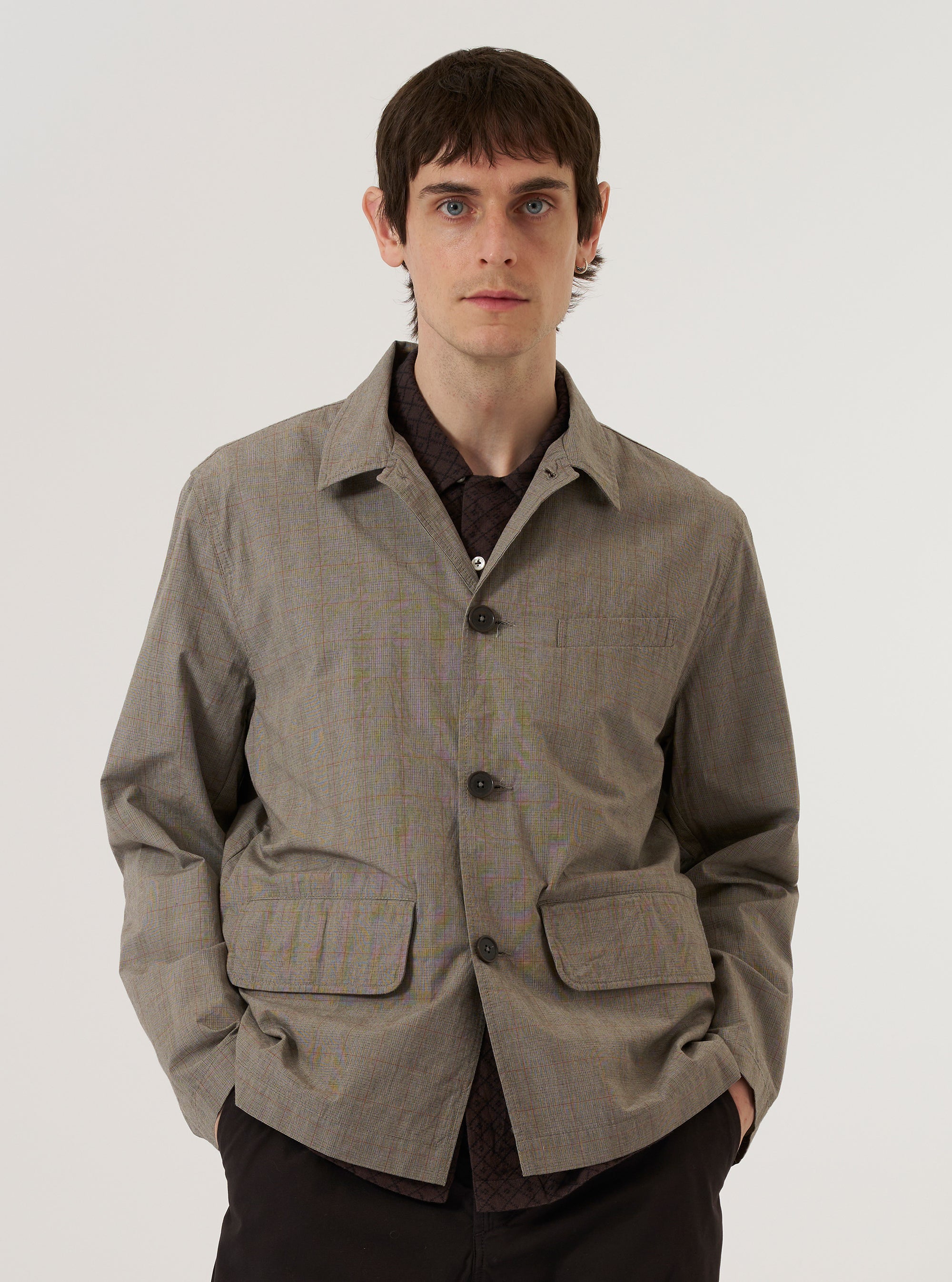 A model wears the Grey Brown Wales Glen Check Franck Jacket over a light-colored top, standing against a plain white background. The jacket features a shorter cut, a one-piece flat collar worn open, and two large front flap pockets. The fabric displays a detailed grey and brown glen check pattern. The model is shown from the front, highlighting the garment's workwear-inspired silhouette and button-down closure.